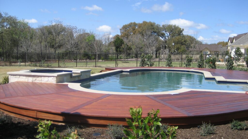 Raised wood deck with round inground pool, spa, and black fencing in large backyard.