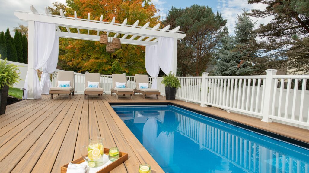 Rectangular pool deck with white pergola, lounge chairs, and light wood flooring.