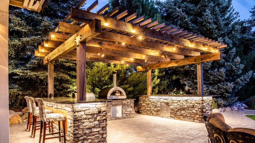 Wooden pergola over stone outdoor kitchen with bar seating, string lights, and pizza oven.