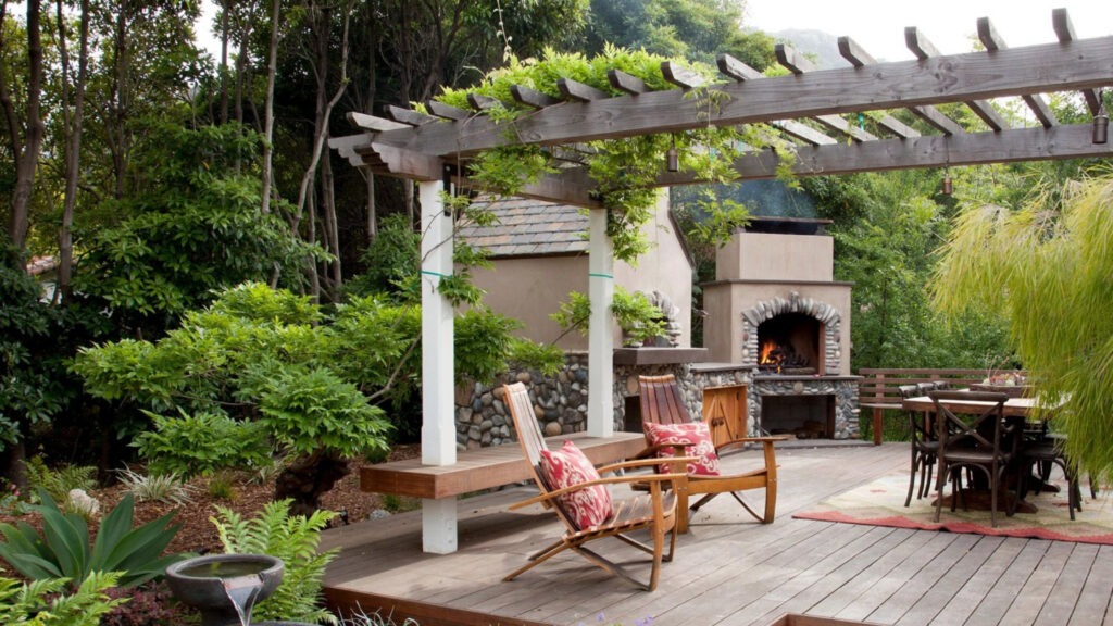 Rustic garden pergola with stone fireplace and wooden chairs in lush setting.