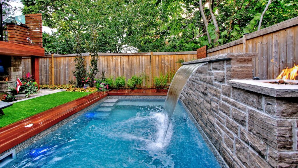 Small backyard pool with wood deck, stone waterfall, and fire feature.