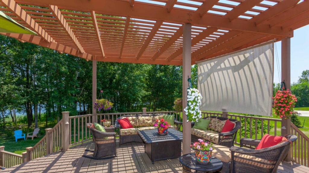 Traditional brown pergola on deck with wicker furniture, flowers, and colorful cushions.