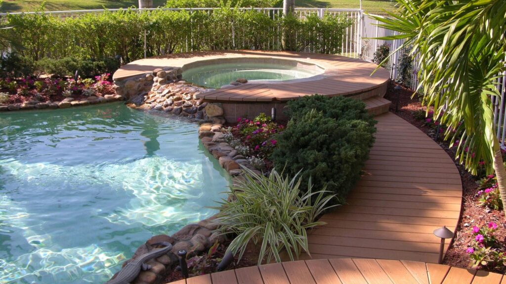 Tropical above ground pool deck with wood pathway, natural rocks, and lush plants.