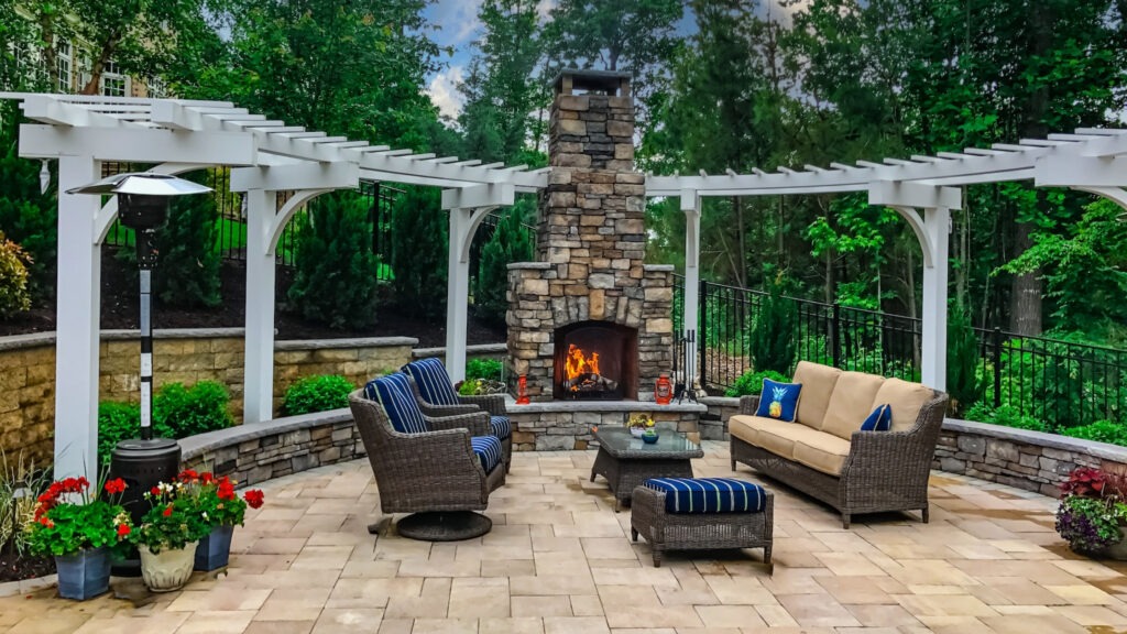 White curved pergola surrounding stone outdoor fireplace with wicker chairs and lush garden.