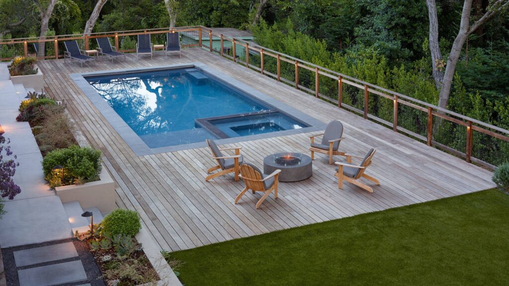 Elevated wood pool deck with firepit, lounge chairs, and glass railing in forest setting.