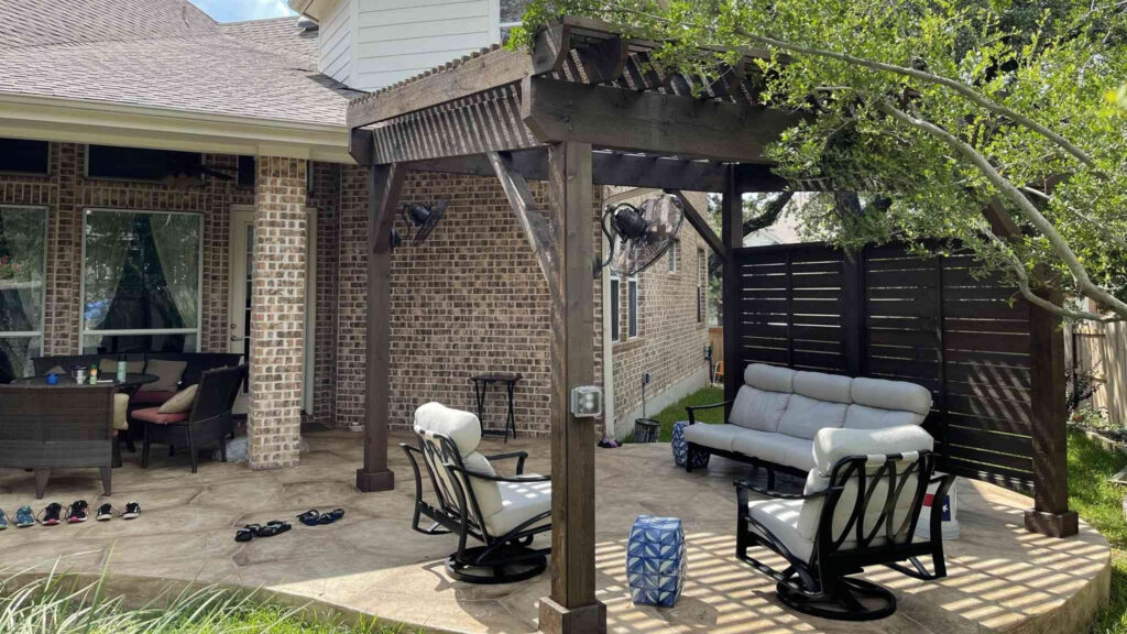 Wooden pergola attached to brick house with cushioned seating and privacy wall.