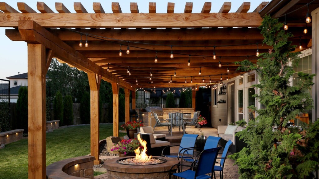Wooden pergola with hanging lights, stone firepit, and outdoor seating area on backyard patio.