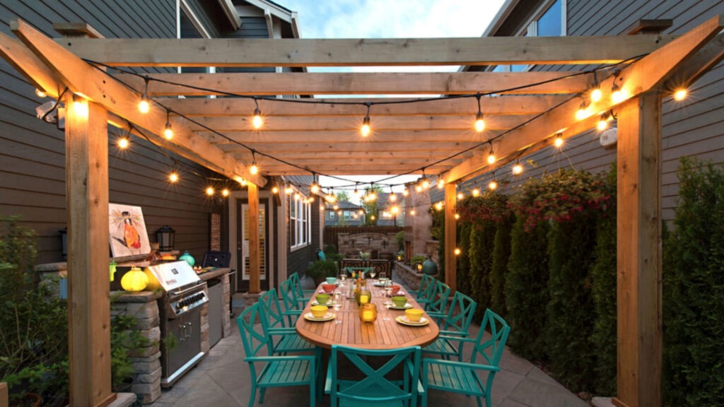 Wooden pergola with hanging string lights, teal dining chairs, and outdoor grill setup.