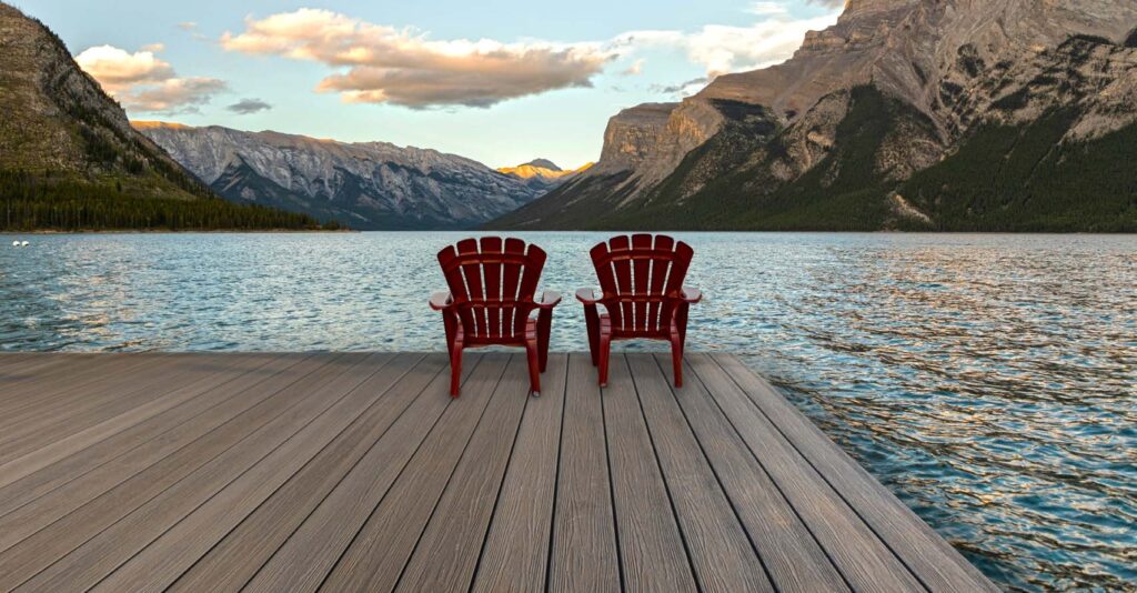 A lakeside dock built with Eva-Last Eclipse Autumn Mahogany decking, set against a backdrop of mountains and calm blue water. Two red Adirondack chairs face the lake, and the rich brown deck boards show subtle woodgrain detail that blends naturally with the outdoor scenery.