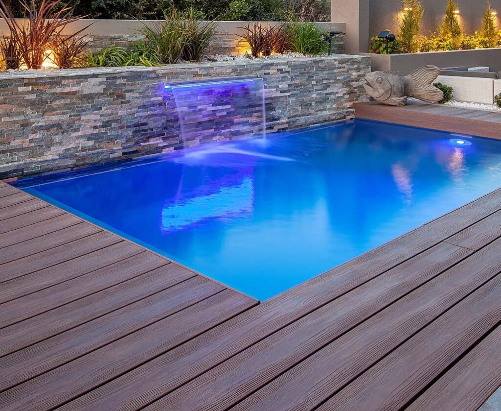 A modern poolside deck made with Eva-Last Eclipse Autumn Mahogany composite decking, featuring a smooth woodgrain surface beside a bright blue pool. A stone wall with a glowing water feature adds ambiance, showcasing how the decking resists moisture and complements outdoor luxury settings.