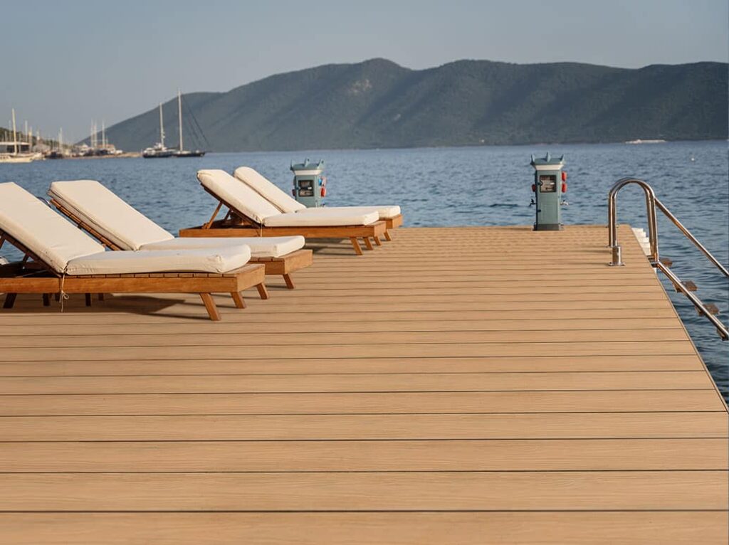 A serene dockside setting featuring Eva-Last Eclipse Dockside Brown decking, paired with cushioned wooden lounge chairs overlooking calm blue water and distant green hills under a clear sky.