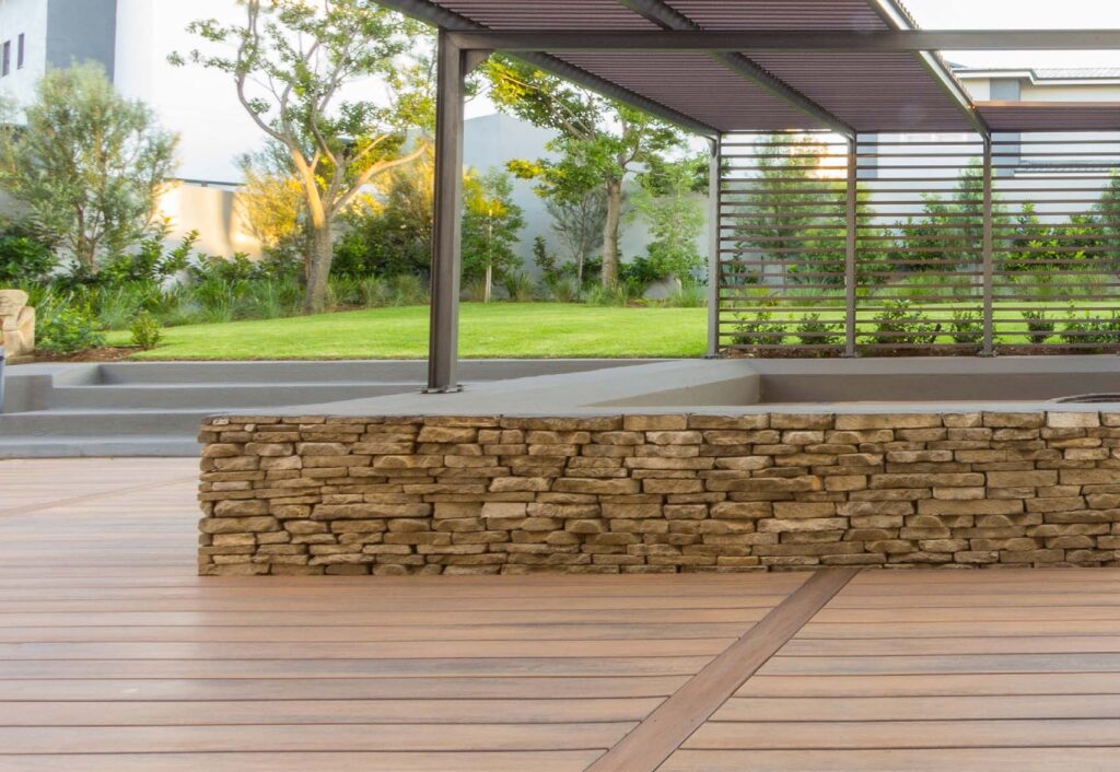 A warm garden-style patio featuring Eva-Last Eclipse Dockside Brown decking, a stacked stone wall, modern pergola structure, and a landscaped yard with trees and shrubs in the background.