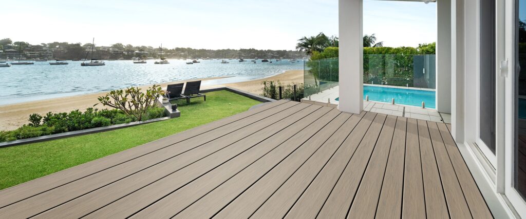 An outdoor deck built with Eva-Last Eclipse Shoreline Gray boards overlooking a calm waterfront with boats anchored in the distance. The soft gray planks show subtle woodgrain detail and create a smooth, modern surface that blends naturally with the coastal landscape. The deck leads to a grassy lawn and pool area beneath a bright, clear sky.