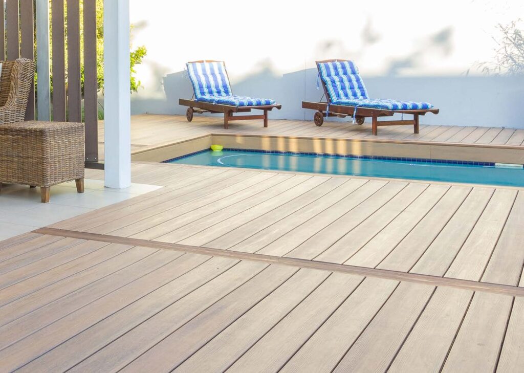 A backyard pool area featuring Eva-Last Eclipse Shoreline Gray composite decking with its pale, driftwood-like color and textured grain. Blue-striped lounge chairs sit on the elevated deck while the lower section wraps neatly around the pool edge, highlighting the slip-resistant, moisture-safe design ideal for wet environments.