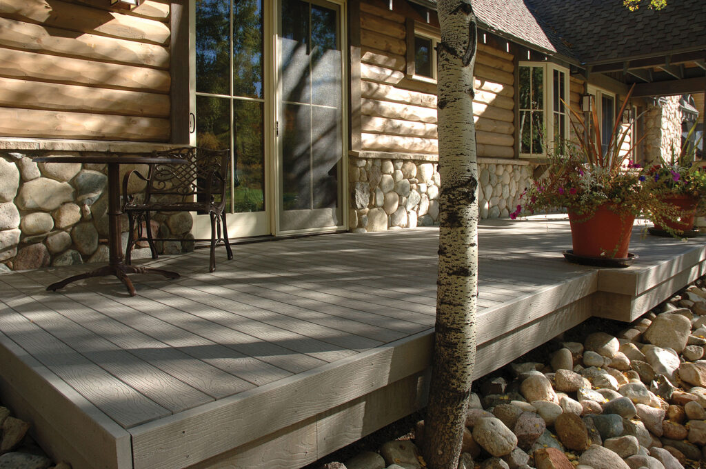 Envision EverGrain Cape Cod Grey composite decking surrounding a rustic cabin, paired with stone accents and natural landscaping. The soft gray color and deep grain pattern complement the wood exterior, creating a cohesive, natural-looking outdoor living space.