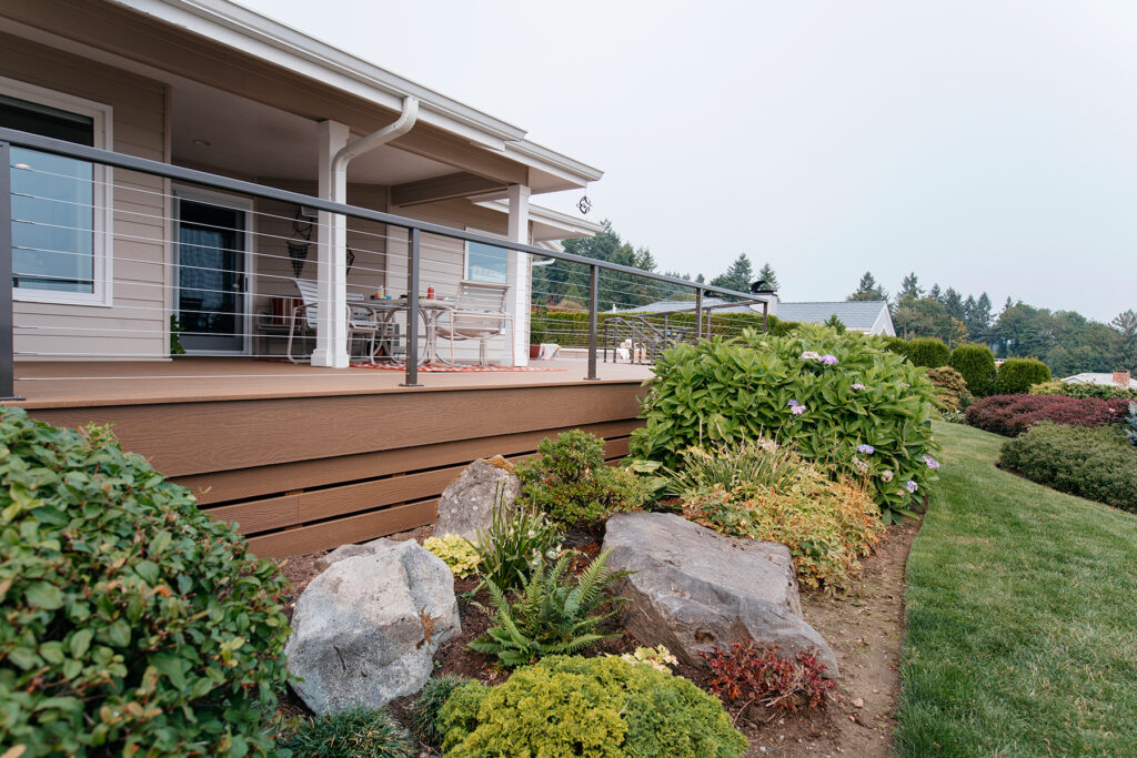 Weathered Wood composite decking shown on an elevated backyard deck with modern cable railing and clean trim details. The warm, muted brown tones create a natural transition between the home exterior and landscaped greenery below. The deck surface appears smooth and consistent, offering a balanced, understated wood look suited for everyday outdoor living.