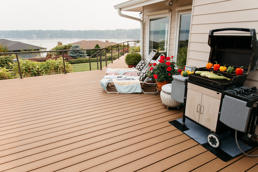 Weathered Wood composite decking used on a spacious outdoor deck featuring a grill station, lounge seating, and lake views in the distance. The soft brown color adds warmth without overpowering the space, complementing light siding and metal railing systems. The decking boards run cleanly across the surface, highlighting a refined, low-maintenance outdoor gathering area.