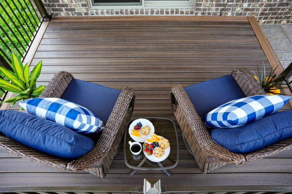 Outdoor deck finished in Envision Ridge Premium Black Walnut composite decking, featuring deep brown tones with subtle woodgrain variation. The deck is styled with woven lounge chairs and blue cushions, creating a warm, inviting seating area. The rich color adds contrast against the home’s brick exterior and surrounding greenery.