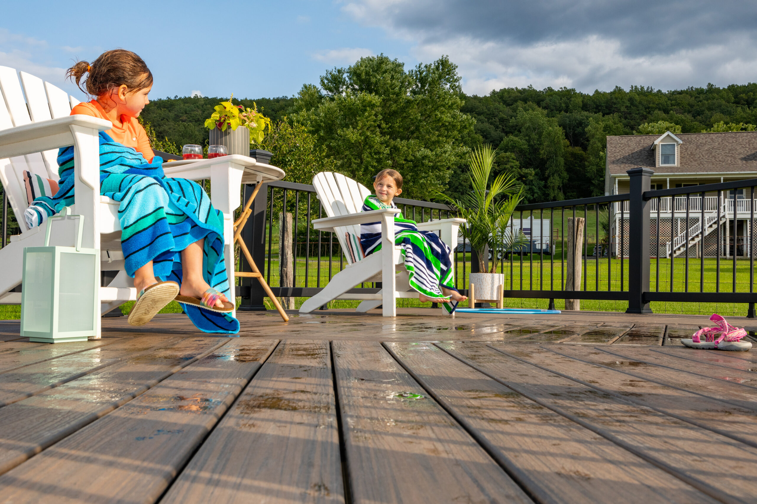Two children sit on white deck chairs on a wet composite deck after swimming, with water visible across the deck surface. This image shows how outdoor spaces often stay damp after use, which leads homeowners to ask, is Trex decking slippery when wet, especially in family settings.