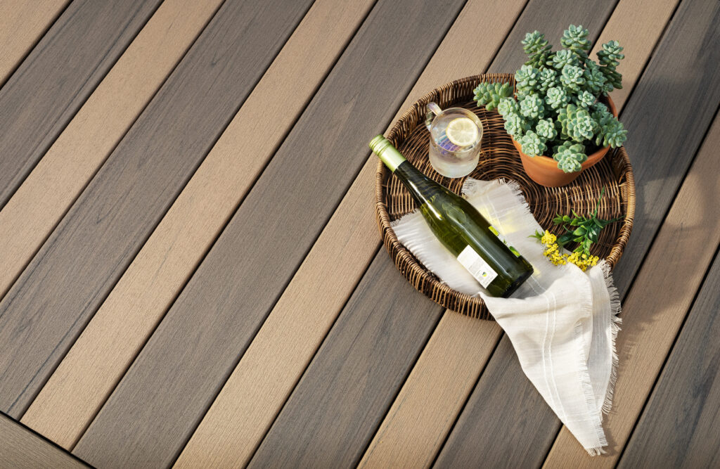 Composite decking boards with natural wood-style tones, shown with a decorative tray, plant, and bottle. The capped surface highlights why paint or deck stain does not absorb into composite materials.