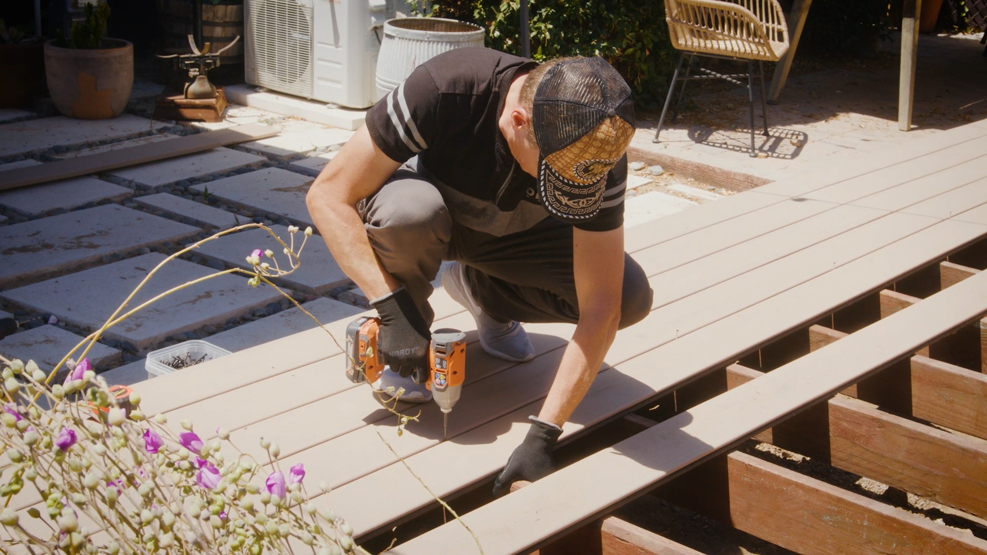 Contractor securing composite deck boards to a wood deck frame using a drill. The boards run perpendicular to evenly spaced joists set at standard composite decking 16 inches on center spacing.