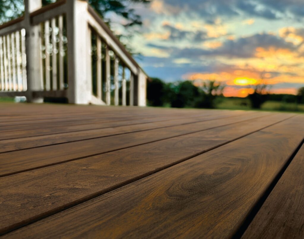 Fiberon Novus Natural Ipe composite decking installed on a residential deck overlooking a sunset landscape. The warm brown woodgrain surface enhances the white railing system and creates a refined, high-end outdoor living space.