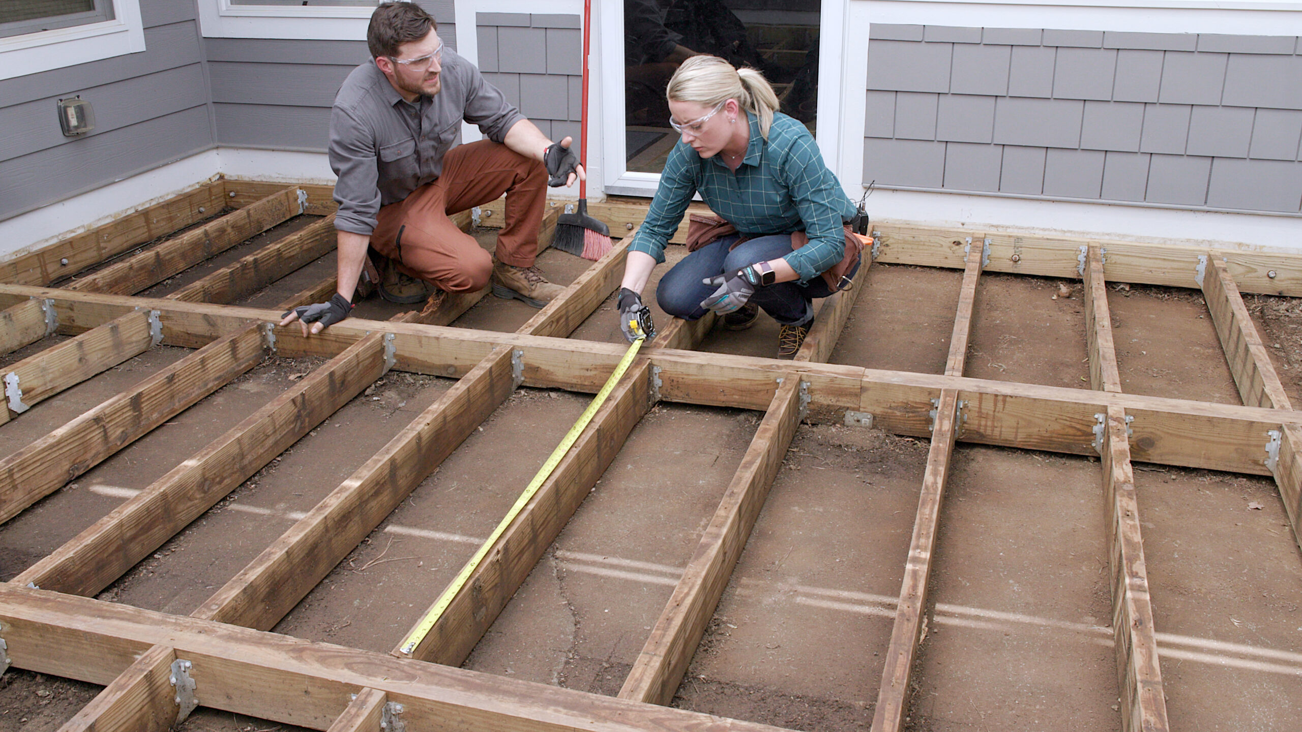 Two contractors measuring joist spacing on a wood deck frame before installing composite decking. The image shows evenly spaced treated wood joists attached with joist hangers, demonstrating proper deck frame layout and decking joist spacing preparation.