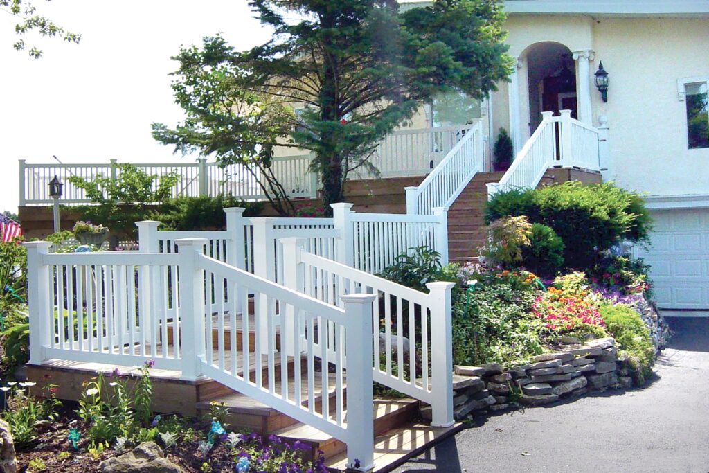 Westbury Cardinal vinyl railing installed on a multi-level front entry deck with stairs, featuring white posts and balusters surrounding landscaped garden beds and a residential home exterior.
