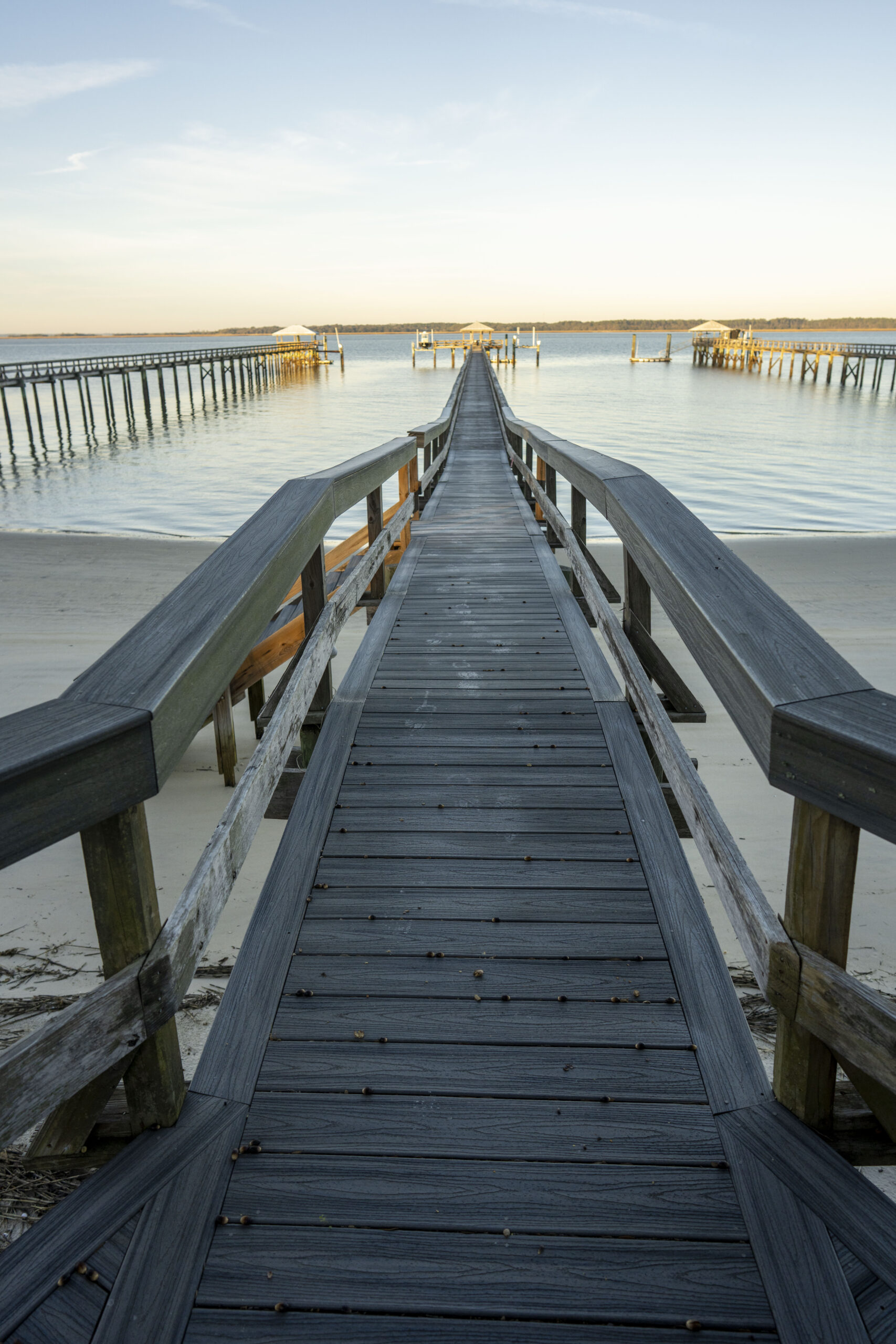 Wooden boardwalk pier extending over shallow coastal water toward covered dock platforms in the distance