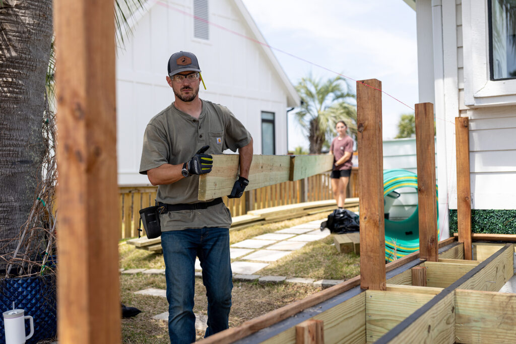 Builder carrying ProWood treated lumber past a low deck frame with 4x4 posts and a string line set.