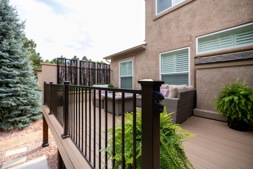 Bronze Envision Fairway modern steel railing with a lit post cap on a tan composite deck beside a stucco home, warm metal deck railing ideas.