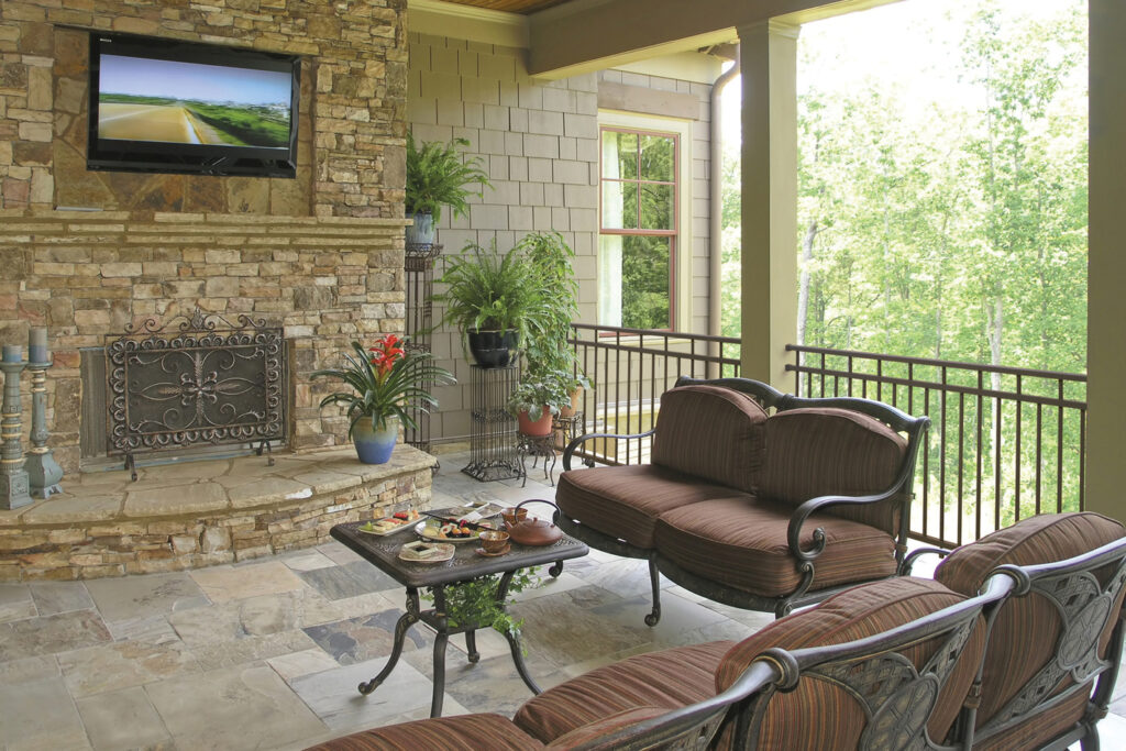 Textured bronze Westbury Riviera aluminum railing on a covered porch with a stone fireplace and wooded view, warm metal deck railing ideas.