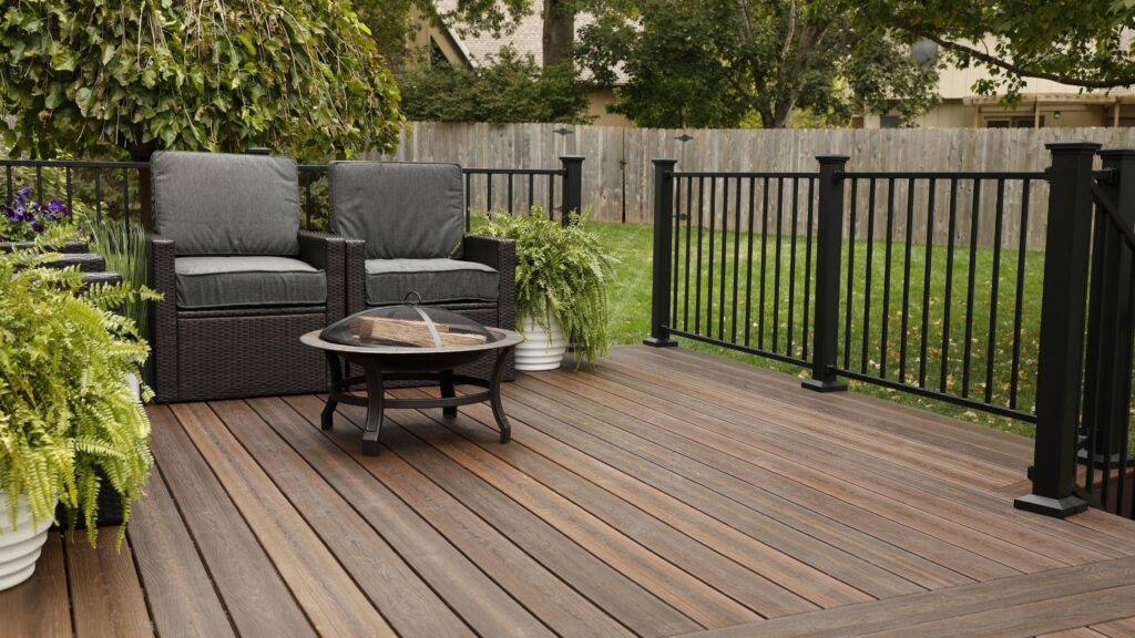 Envision deck ideas featuring Distinction Rustic Walnut decking with black railing, cushioned chairs, fire pit, and potted ferns.