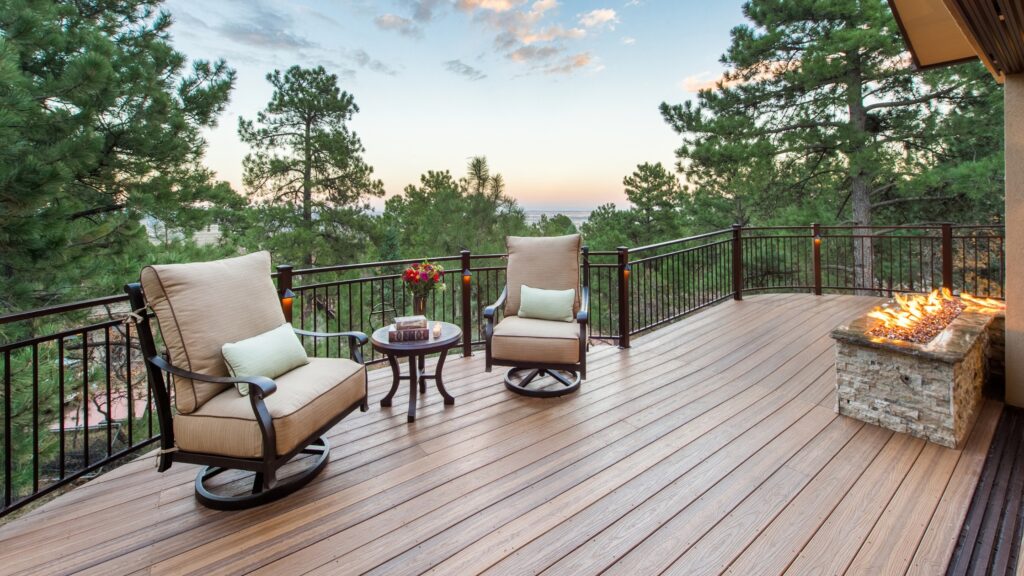 Envision Distinction Spiced Teak deck with black railing, cushioned chairs, stone fire feature, and pine tree views.