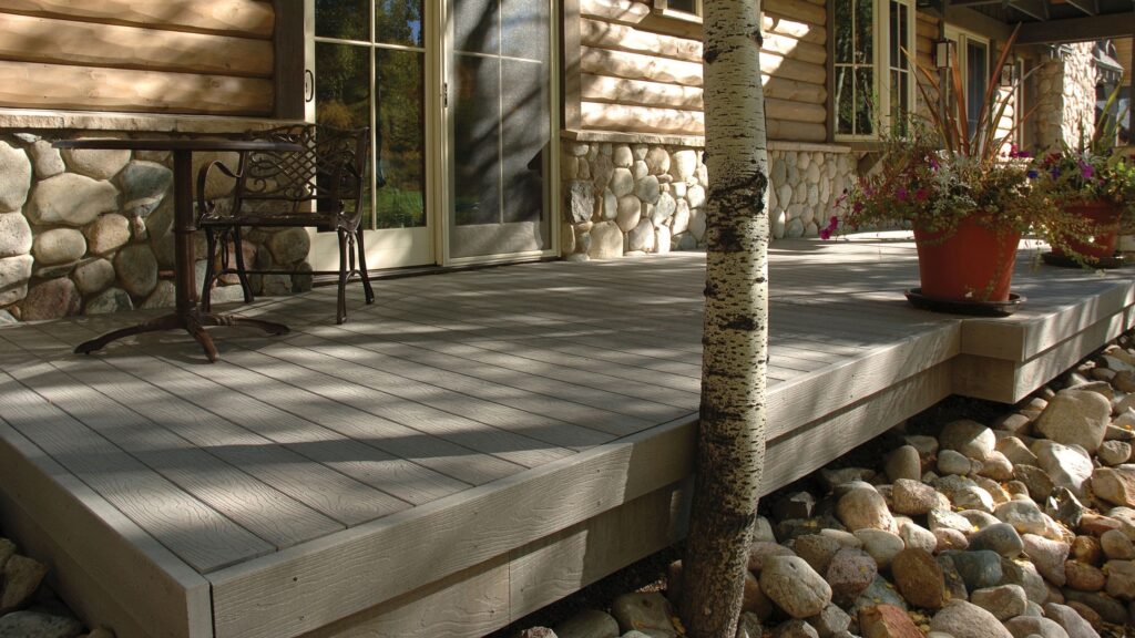 Envision deck ideas featuring EverGrain Cape Cod Grey decking beside a rustic log home with stonework, river rock, and a mature tree.