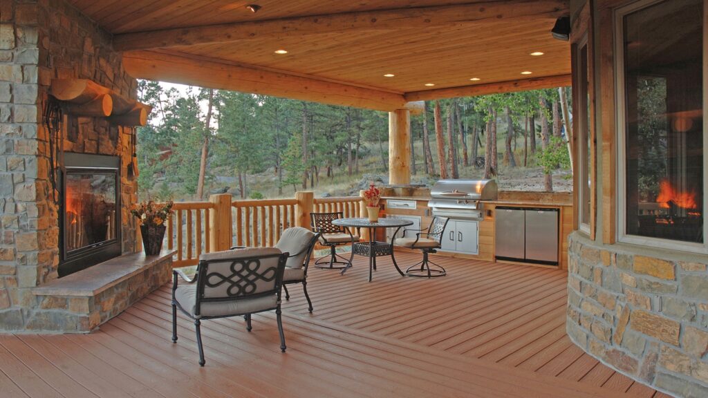 Envision EverGrain Redwood deck with stone fireplace, built-in outdoor kitchen, log railing, dining furniture, and covered timber roof.