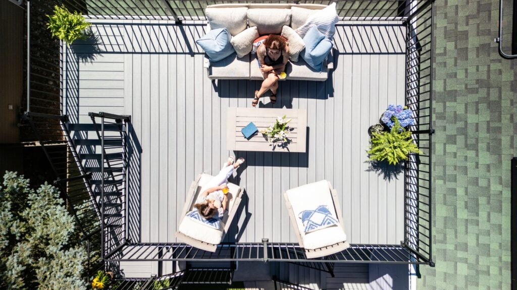 Envision Ridge Premium Alpine Fog deck with sofa, chaise lounges, central coffee table, black railing, and flowering planters.