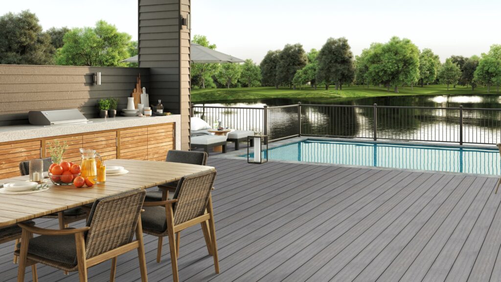 Envision Ridge Premium Alpine Fog pool deck with outdoor kitchen, dining area, black railing, and waterfront landscape.
