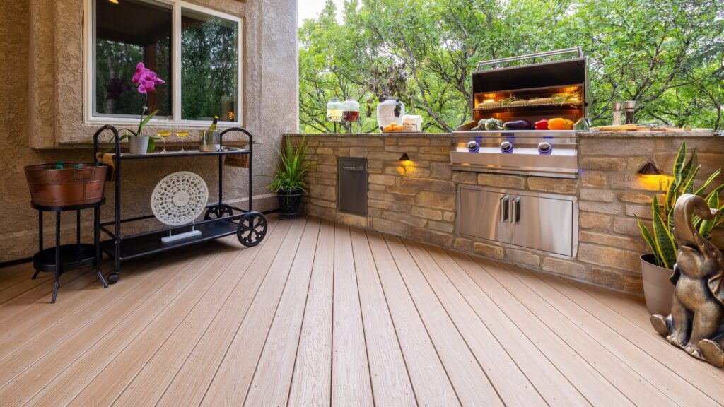 Envision deck ideas featuring Ridge Premium Sandstone decking with a stone outdoor kitchen, built-in stainless steel grill, serving cart, and wooded surroundings.